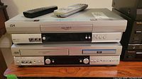 Front stacked view showing a silver JVC DVD/VHS combo player on bottom and VHS player on top with remotes on top of the VHS player.