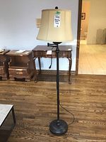 Full view of floor lamp showing metal base, black metal pole, beige fabric shade, and attached power cord lying on hardwood floor.