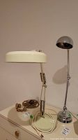 Both lamps shown side by side, including the cream-colored magnifying lamp by Luxo and the chrome adjustable desk lamp by AEL Lighting Co on a cream-colored surface.