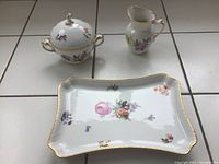All three items displayed sitting on white tile, showing the set together: lidded sugar bowl, creamer, and rectangular platter decorated with flowers and gold trim.
