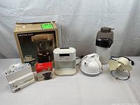 Seven assorted small appliances arranged on a white surface against a white background. Includes boxed Proctor-Silex coffee maker, electric can opener in box, white kettle, pop corn machine, fan, and others.