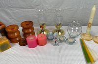 Full view of assortment of candles and candle holders arranged on white cloth background including wooden candle holders, glass hurricane candle holders, glass votive holder, pink square candles, white taper candles, and assorted colored taper candles.