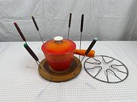 Orange ceramic fondue pot placed on round wooden holder, surrounded by eight fondue forks with wooden handles, with metal warming stand beside it.