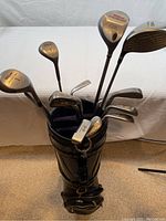 Photo showing a black golf bag filled with a set of golf clubs including 3 woods, irons, wedges, and a putter visible on the top in a room with white backdrop.