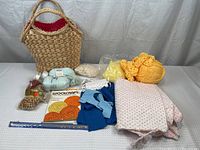 Wicker basket handbag with various knitting and crafting supplies including wool yarn, ribbons, quilting fabric, needles, macrame hook, wooden beads, and orange woven coasters.