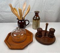 Photo showing MCM kitchen items including a wooden cheese tray with glass dome, wooden salt and pepper shakers on a base, a pottery vase, and a ceramic jug holding wooden utensils.