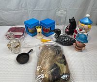 Photo showing assortment of ceramic piggy banks, glass jar with lid, black cat and loon figurines, plastic dog, frying pan, ladle, and costume wig bag