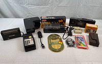 Wide shot of the lot showing all items including radios, battery charger box, camera box, pedometer, reading light, phone case, cables, and other small items arranged on a white surface.