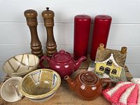 Photo showing all items in the lot arranged on a wooden surface: two ceramic teapots, two pottery bowls, wooden salt and pepper mills, two large red pillar candles, a ceramic decorative house, and a ceramic bell.