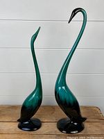 Photo showing two ceramic geese figurines on a wooden surface against a white shiplap background. Both have teal to black gradient glaze with long slender necks.