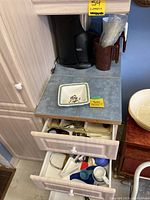 Coffee maker on countertop with brown measuring cup and clear plastic cups beside, white and green Portmeirion square bowl in front, plus two drawers open showing assorted kitchen tools and gadgets.
