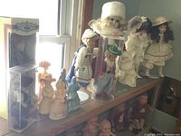 Shelf showing assorted porcelain and plastic dolls dressed mainly in vintage style clothes, including bride and Dutch dolls, some with hats or bonnets.
