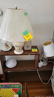 Three lamps on wooden shelf: ceramic with floral pattern with shade, rectangular white lamp, milk glass hobnail lamp base without shade, two extra glass shades