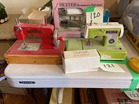 Two miniature sewing machines displayed on a plastic table, including the Kay-Ee Sew Master and Sewmate, with boxes, tags, and visible wear.
