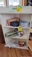 Wide view photo of the small white wooden corner shelf with various medical items placed on three shelves including a green medical case, digital blood pressure reader, boxes of gloves, and pill dispenser.