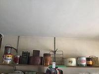 Top shelf containing a line of vintage tins, thermos, pie rack and containers showing wear and age.