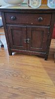 Front view of wooden washstand showing one drawer with two round knobs and double door cabinet below with metal knocker handles. Surfaces show heavy wear.