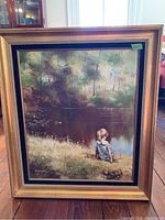 Full front view of the girl at waters edge painting in a gilded gold frame showing the painted landscape and girl sitting at a water body edge with a basket beside her.