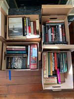 Four cardboard boxes filled with a variety of books on wartime, politics, autobiographies, and hunting.