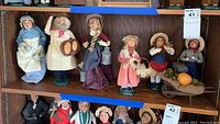 Shelf display of seven Byers Choice caroler dolls in Victorian winter clothing with wheelbarrow containing pumpkin and moss.