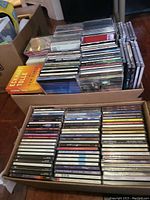 Photo showing two boxes of assorted music CDs stacked inside, with some titles visible on the spines of the CD cases.