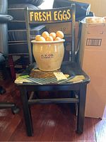 Wooden chair with 'FRESH EGGS' sign on backrest, crock holding faux eggs on seat.