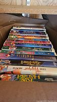 Photo showing a collection of Disney VHS tapes stacked in a box, titles visible include Lady and the Tramp, The Fox and the Hound, Fantasia, Dumbo, and others.