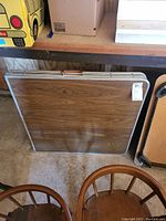 Folding table folded and leaning against a shelf, showing rectangular shape with wood texture and metal edges.