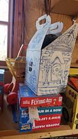 Stack of four boxed board games on wooden surface including Snoopy Flying Ace, Four in a Row, Who's Left?, and Popper Magic games, with a yellow vintage plastic child’s shopping cart in background.