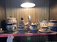 All pieces arranged on glass shelf under light: tureen, Christmas tree, basket, two lidded vessels, cup and saucer, mirror tray