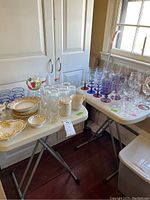 Overview of Lenox mugs, small plates, assorted glassware including wine glasses with colored stems on folding tables.