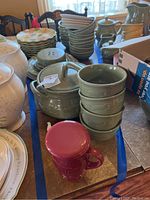 Full view of the assortment of Longaberger dinnerware on a table including sunflower luncheon plates stacked, sage green bowls and tureen, paprika mug with lid.
