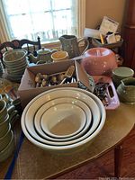 Entire lot arranged on table: green nested mixing bowls, boxed silverware, pink server, pitchers and crocks