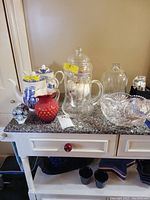 Overview of various pitchers, figurine, two Staffordshire tea pots, cut glass bowl, decanter and glass dome displayed on a granite surface.