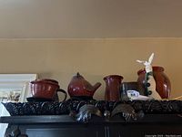 Shelf displaying six pottery items: a ribbed bowl or crock, bird-shaped pitcher, red vase, floral painted pitcher, redware bowl, and bunny figurine.