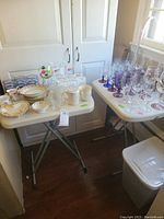Three folding tables sized about 33x28 inches when open, holding multiple glasses, mugs, and small dishware items.