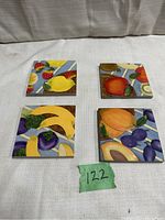 Four separate ceramic square coasters with fruit designs arranged in two rows against fabric background