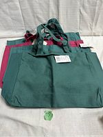 Four canvas tote bags stacked with handle straps visible; three hunter green and one dark pink bag. Tags attached to bags visible indicating new condition.