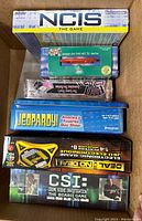 Top-view photo of six factory sealed board games stacked inside a box. Visible titles include NCIS The Game, Bunco, Jeopardy!, Deal or No Deal Electronic Game, and CSI The Board Game.