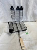 Full view of the black metal BBQ grill cage with wooden handle extended and the cage standing upright showing three long compartments created by dividers.