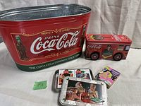 Photo of all items in the lot showing large Coca-Cola metal tin bucket, red truck piggybank, Coca-Cola tin with playing cards, and single pack of playing cards