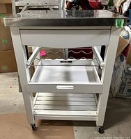 Front view of compact kitchen trolley showing white frame, steel top, wine bottle holder basket, two lower shelves and caster wheels.
