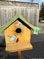 Front view of the wooden birdhouse showing circular entrance, perch, and painted bird design with green roof.
