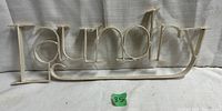 Front view of white metal laundry sign showing the word 'Laundry' in capitalized style with a serif font, against a light fabric backdrop.