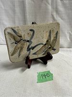 Rectangular speckled stoneware hot plate with abstract black and brown brushstroke design on beige background, displayed upright on wooden stand