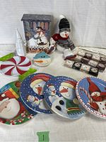 Photo showing mini 2-drawer wood cabinet with snowman family image and Kind Deeds wording, plush snowman figurine, packs of plates, and melamine plates arrayed in front.
