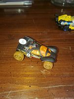 Photo of black off-road style Hot Wheels buggy on a wooden surface, with a yellow and blue Hot Wheels vehicle partially visible in background.