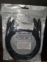 Two black digital optical audio cables in separate resealable plastic bags with connectors visible and labels showing product code, length 1.8m, compatible with sound bar, TV, PS4, Xbox.