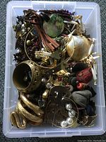 View of a plastic tub filled with assorted vintage jewelry including metal bracelets, bead necklaces, brooches, and other decorative pieces