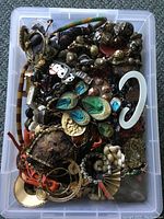 Top view of plastic tub containing assorted vintage and miscellaneous jewelry items including brooches, pins, chains, earrings, and other pieces in multiple colors and materials.
